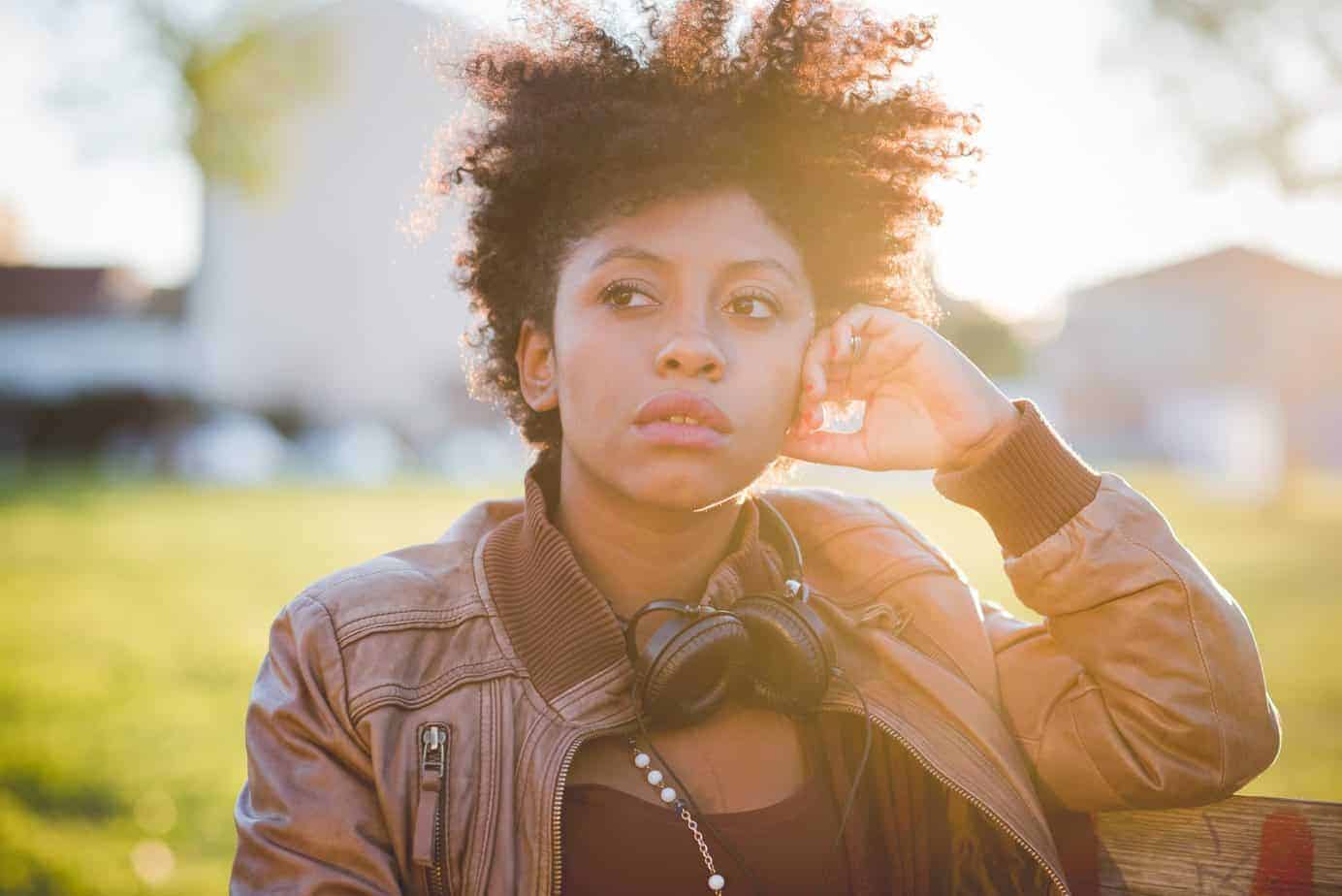 Black girl wearing brown leather jacket with curly hair, standing outside in the sunlight leaning on a fence post. 