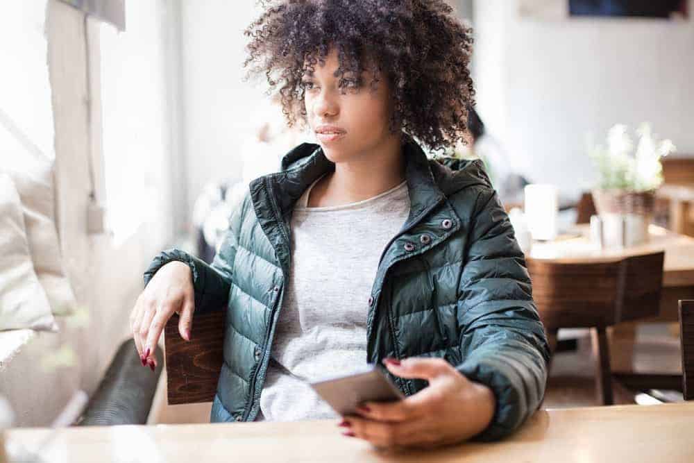 Pretty African American female sitting at a table wearing a green jacket, using a mobile phone with curly hair.