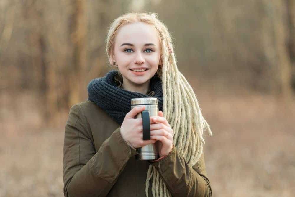 Young beautiful blonde woman with type 1 dreadlocks wearing a scarf and parka posing on a national forest background with thermos coffee cup.