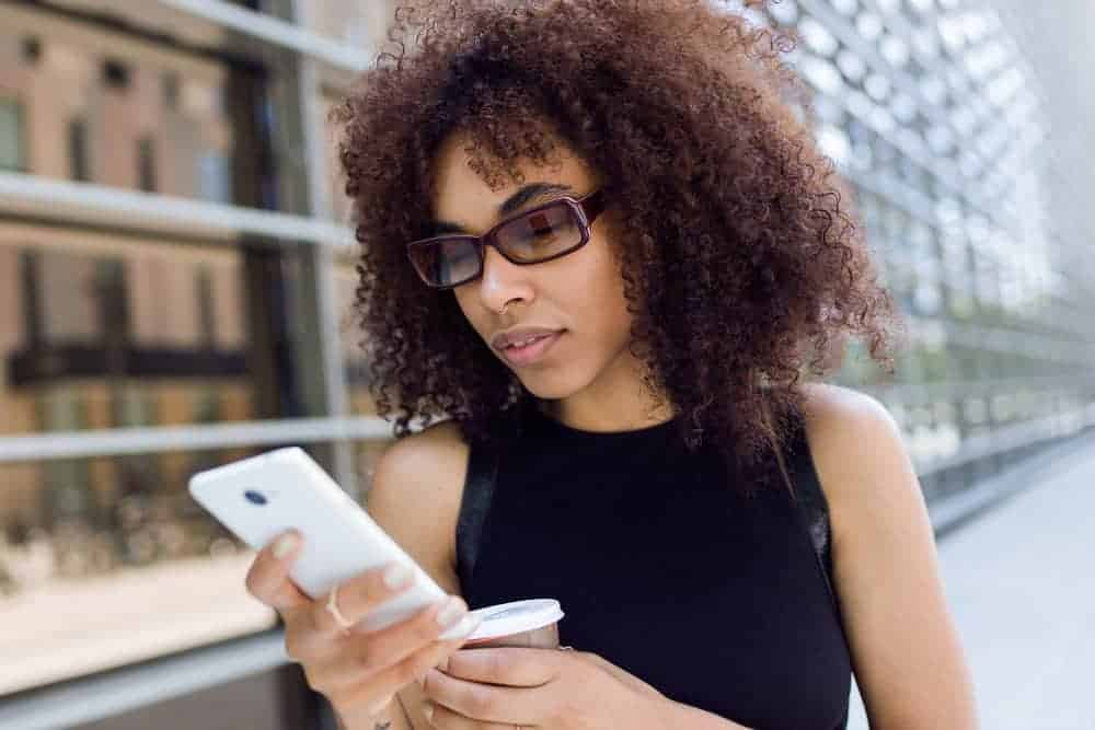 A black female that's using a phone to research the impact that essential fatty acids have on dry or brittle hair strands.