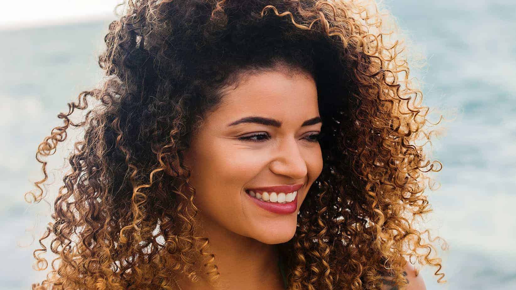 Cute Dominican women with curly dark hair roots and blonde tips smiling on the beach.