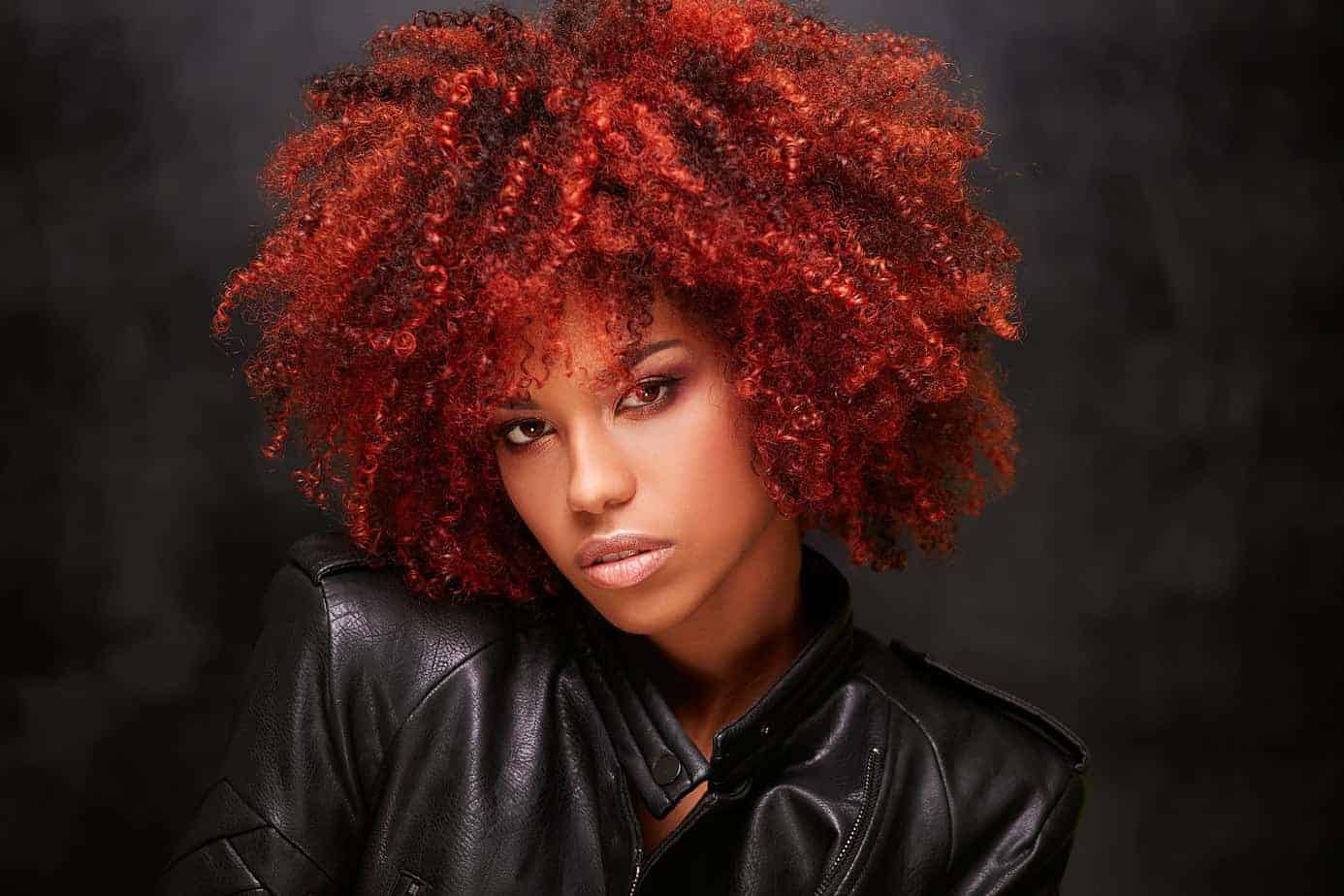 Cute black girl wearing a black motorcycle jacket with red curly hair looking directly into the camera considering the cost of dying her hair again.