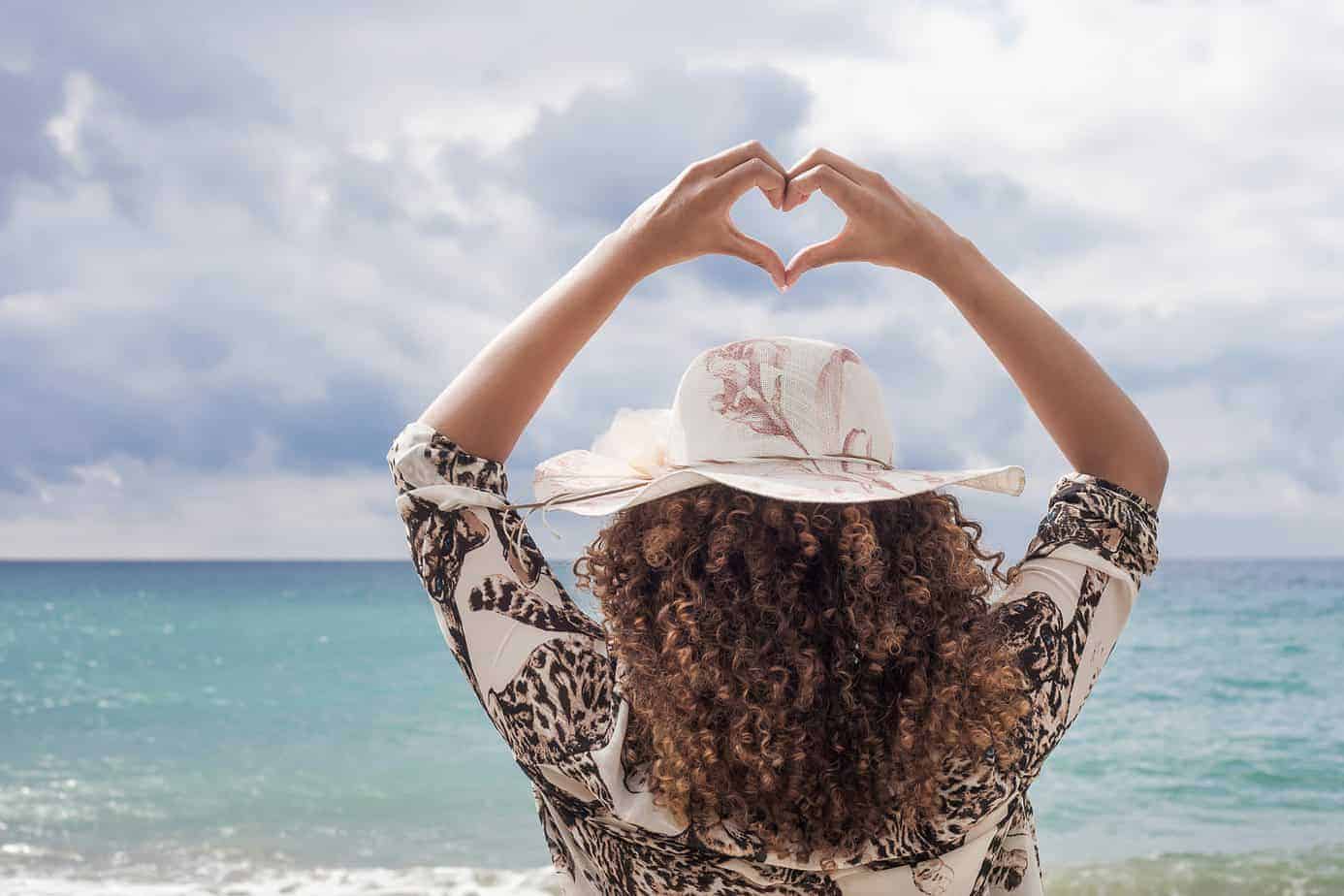 8 Great Benefits of Using Lavender Oil for Hair Care and Growth 9 Woman with curly hair that's been conditioned with lavender oil standing on the beach showing a heart symbol.