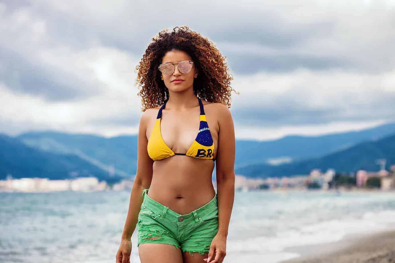 Pretty girl walking on the beach and wearing a yellow bathing suit, green jean shorts, sunglasses and ombre curly hair that's been freshly moisturized with lavender essential oil for hair growth.