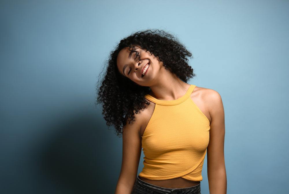 Beautiful African-American woman on blue background wearing a yellow body shirt and blue jeans. Her head in leaning to her right and she has a huge smile on her face, while wearing a wavy hairstyle.
