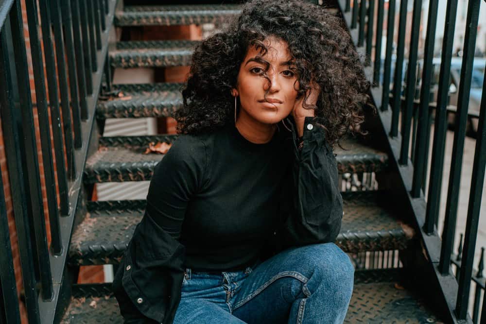 Black women with natural hair treated with sunflower oil sitting on the stairs of an apartment complex, wearing a black mock turtle neck shirt and blue jeans. 