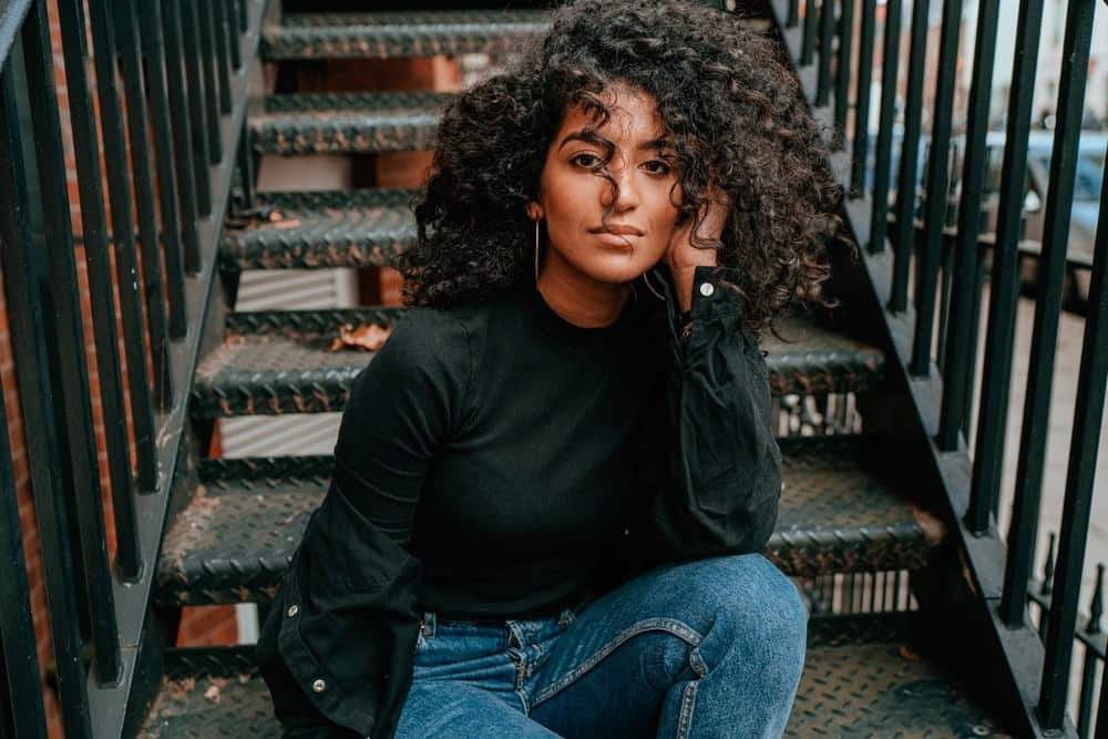 Black women with natural hair treated with sunflower oil sitting on the stairs of an apartment complex, wearing a black mock turtle neck shirt and blue jeans.