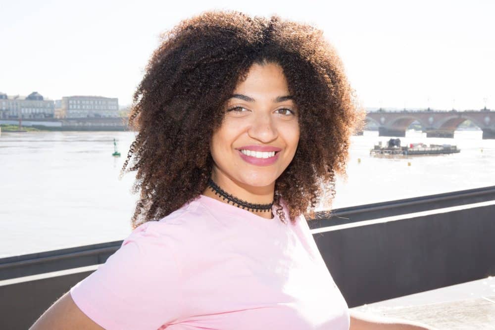 Cute black women smiling standing near river city walk with black necklace, pink t-shirt and ombre curly hair treated with lemongrass oil.