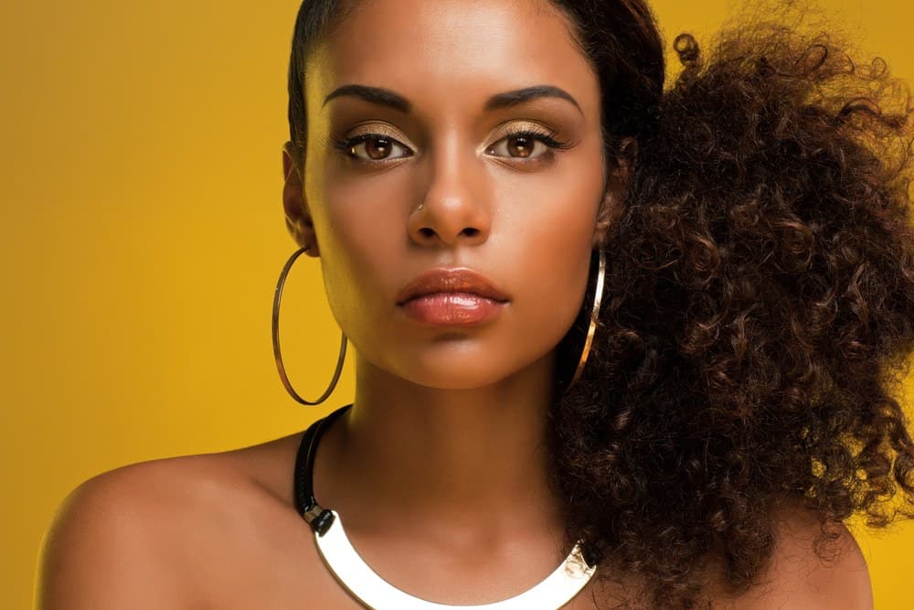 Pretty black women with a curly ponytail hairstyle looking directly into the camera wearing gold ear rings, a nose ring, gold necklace and redish pink lipstick.