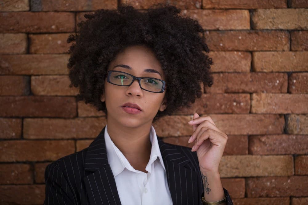 Beautiful African American business woman wearing glasses in the office standing in front of a brick wall.