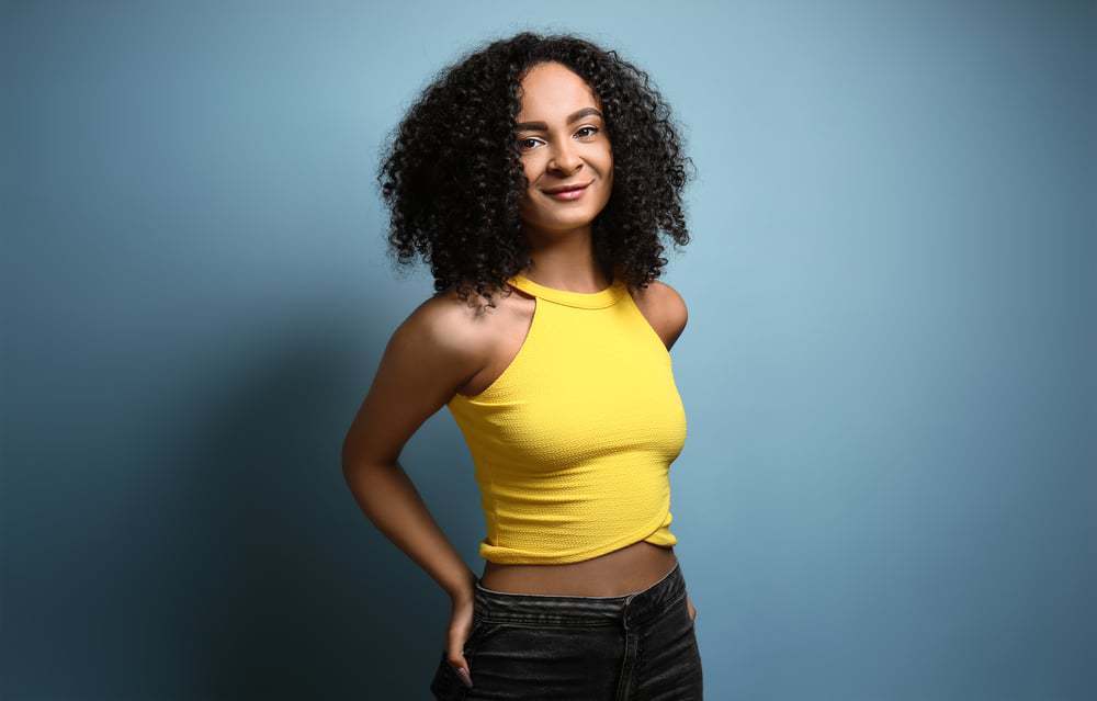 Beautiful African-American woman wearing a yellow shirt and blue jeans with naturally wavy hair that appears to be type 3b.