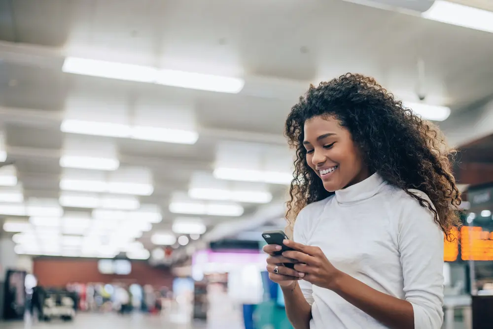 Young woman at the airport, with type 3b hair holding a smartphone and texting wearing a white turtle neck shirt.