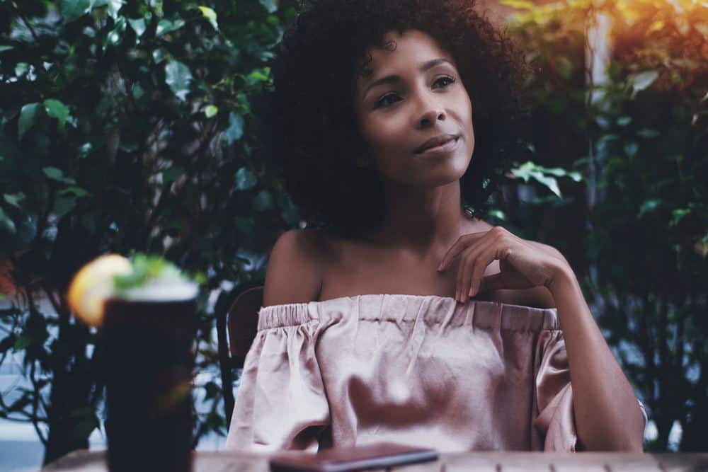 A relaxed curly mixed female is resting in a street bar with a glass of delicious cocktail next to her on the table.