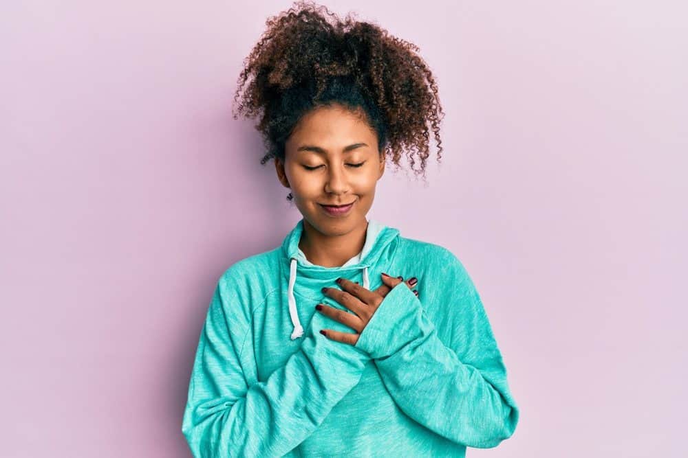 Beautiful African American woman wearing a blue hoodie sweatshirt with her hand over her heart.