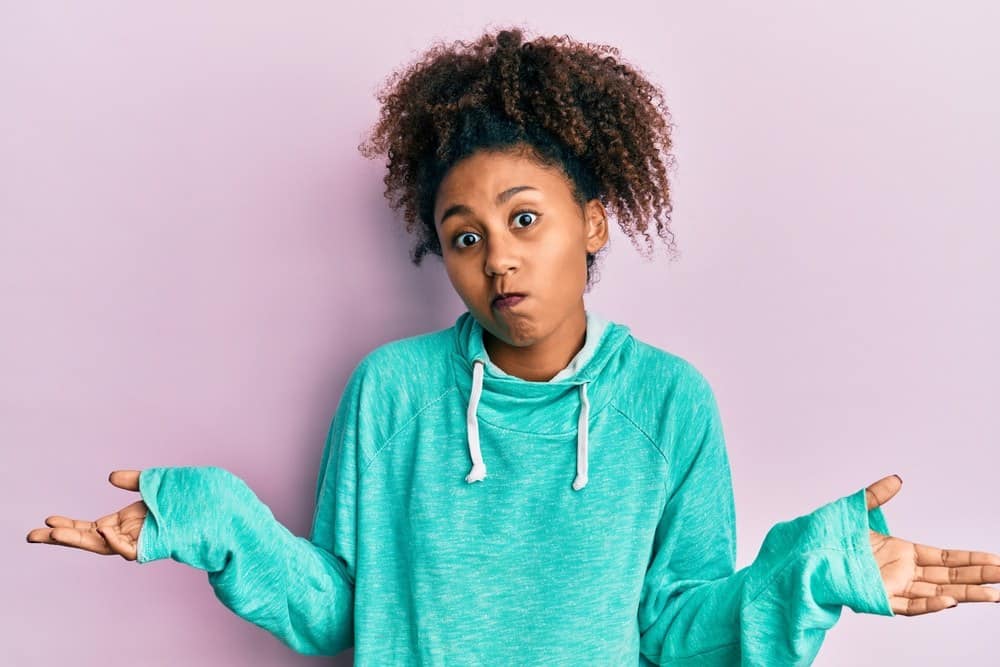 Beautiful African American woman with curly hair wearing casual sweatshirt clueless and confused expression with arms and hands raised.