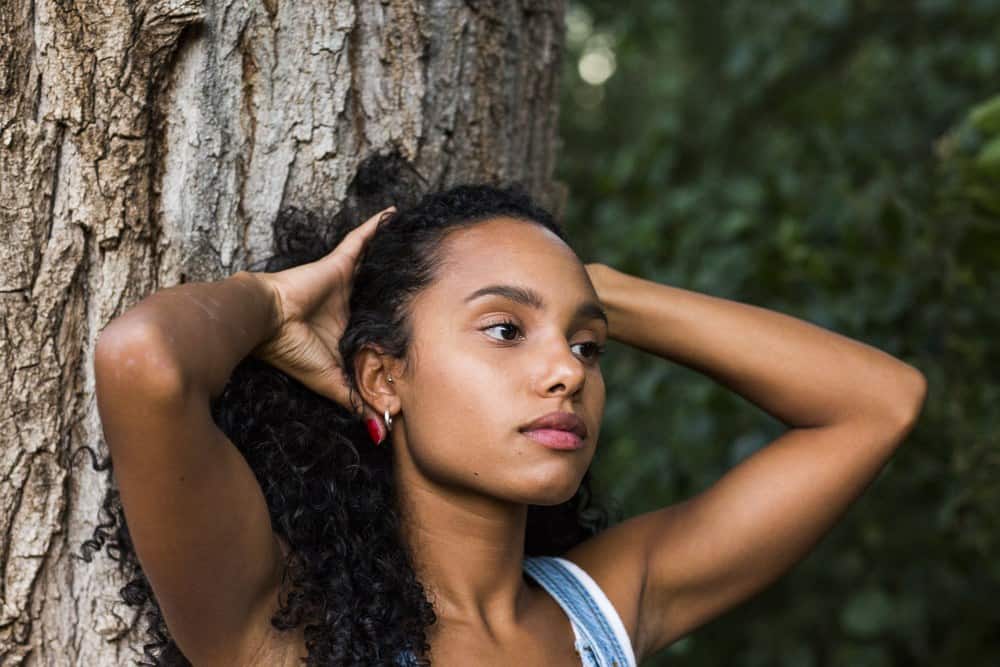 Light skinned biracial women with curly hair standing in front of a tree with her hands creating a ponytail.