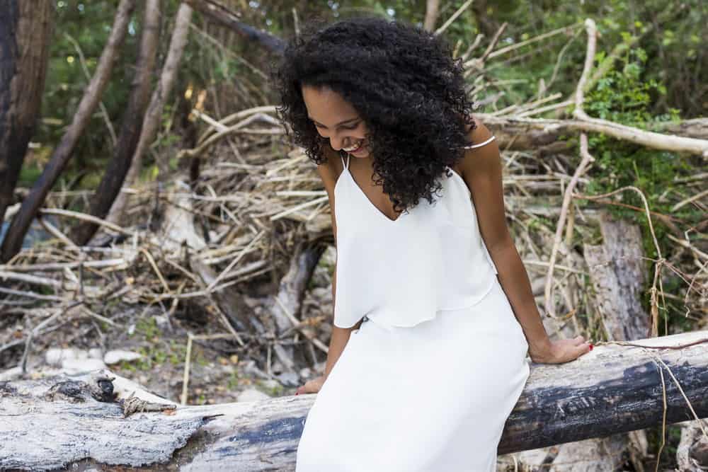 Beautiful young African American woman with clary sage oil treated hair at sitting on tree batch at sunset wearing a white dress.