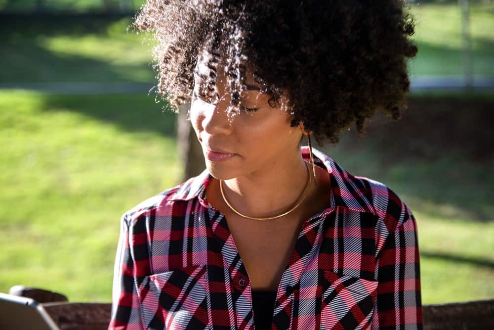 Young beautiful Brazilian female with black curly Afro hair with tablet, while sitting outdoors on the wooden bench in a park