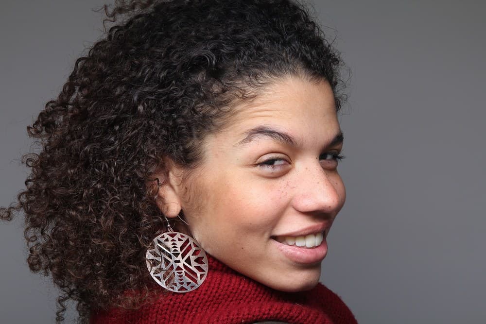 Latino female wearing a red sweater, silver earrings, and ombre type 3c curly hair look back at the photographer while taking a picture.