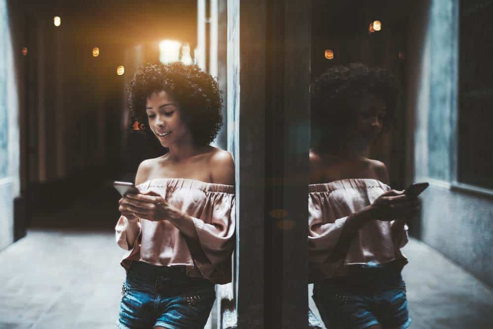 A cheerful young Brazilian female with a medium curly hairstyle is leaning against the wall on a partly dark street corner.