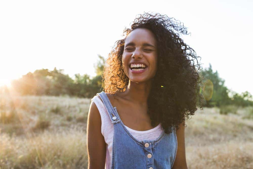 Women wearing blue jean overalls with naturally curly hair that's be moisturized with clary sage oil standing in a field with grass and trees.