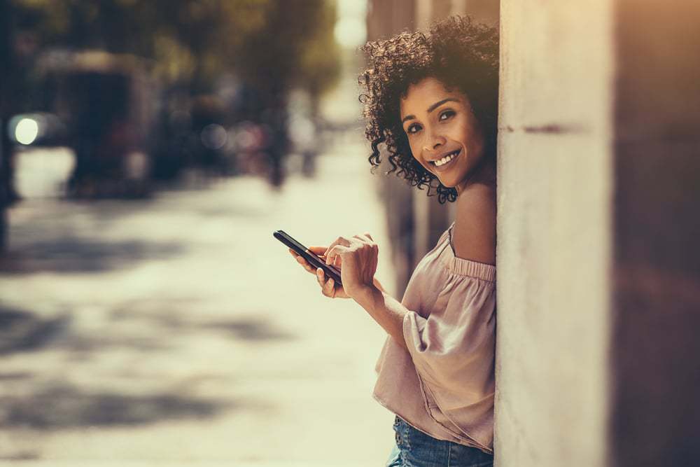 Straw Curls: How to Curl Your Hair with Straws (and Rubber Bands) 5 A young cheerful flirtatious curly-haired biracial female with flexi rods standing outdoors is leaning against the stone column while typing a message using the smartphone.
