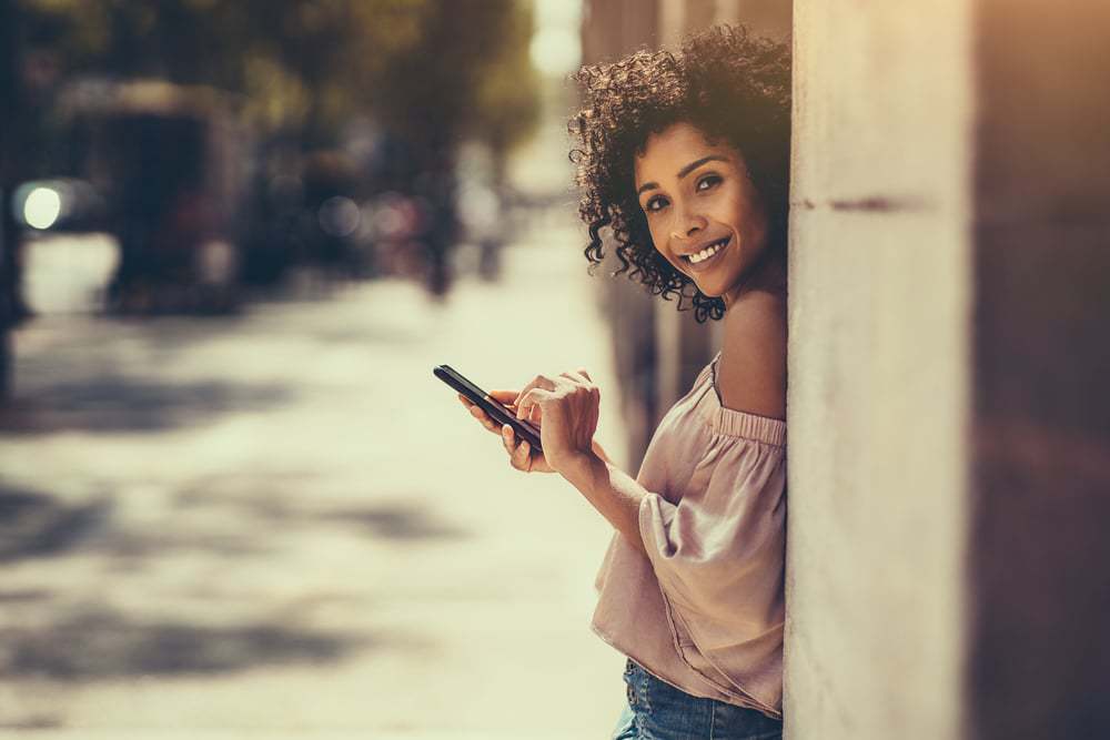 A young cheerful flirtatious curly-haired biracial female with flexi rods standing outdoors is leaning against the stone column while typing a message using the smartphone.