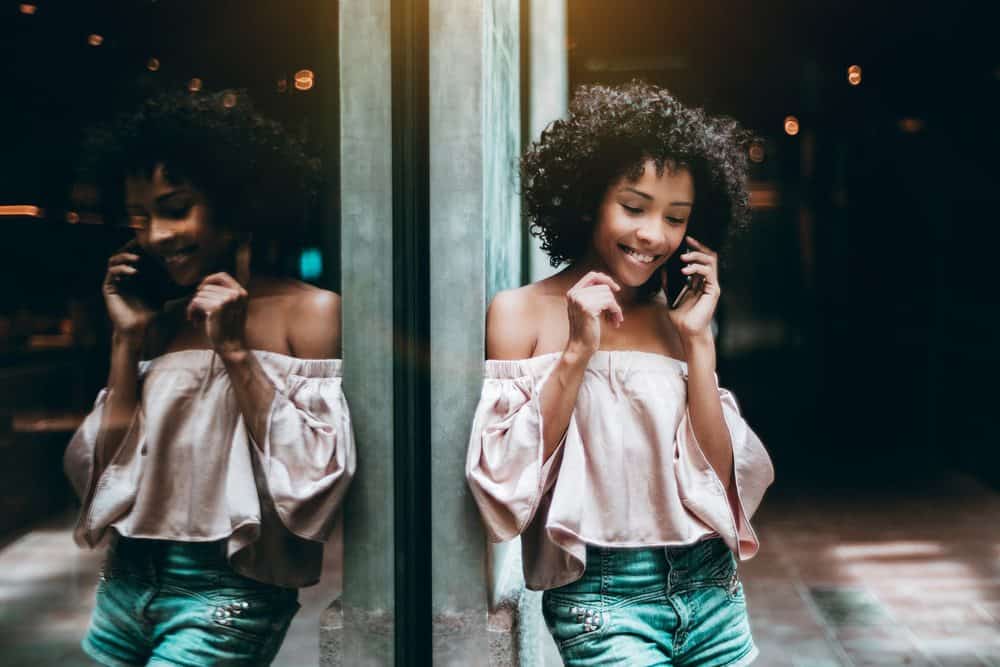 A young African American women with a straw set on type 3c naturally curly hair is having a phone conversation on her iPhone with her boyfriend, smiling while leaning against the wall.