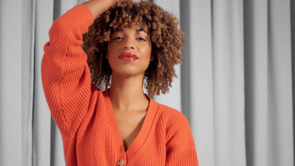 Mixed race black woman with Shirley Temple black textured curly afro hair in bright orange jacket with natural makeup for dark skin tones.