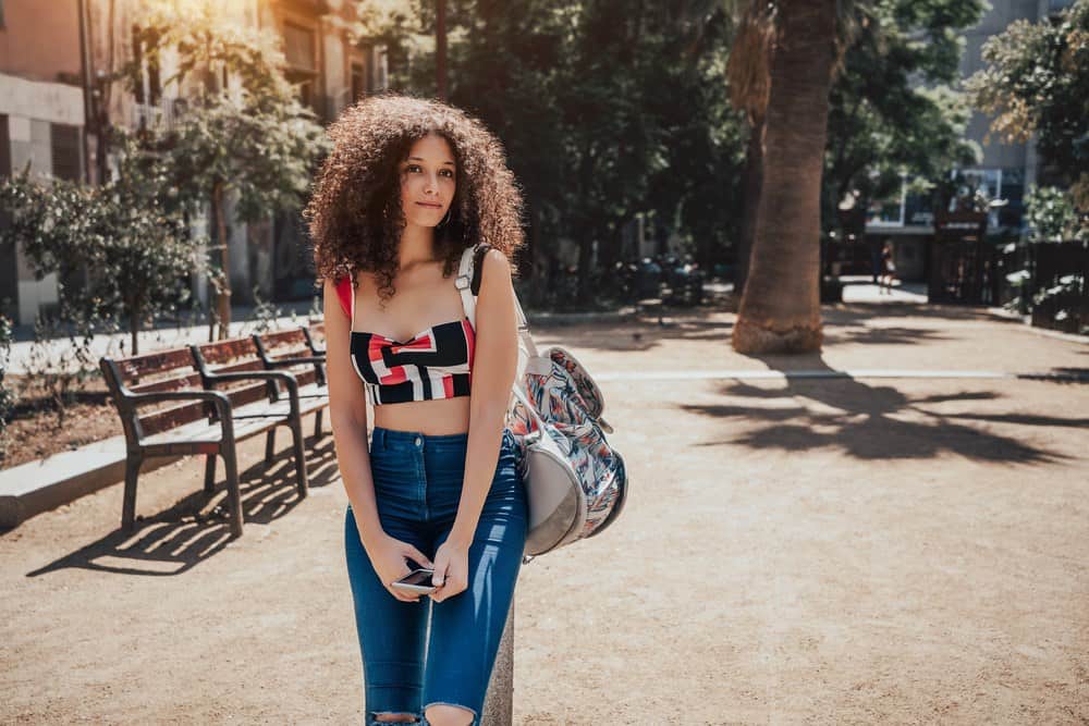 Female in United Kingdom wearing blue jeans, a multi-colored bag, and a red and black crop top with naturally curly hair.