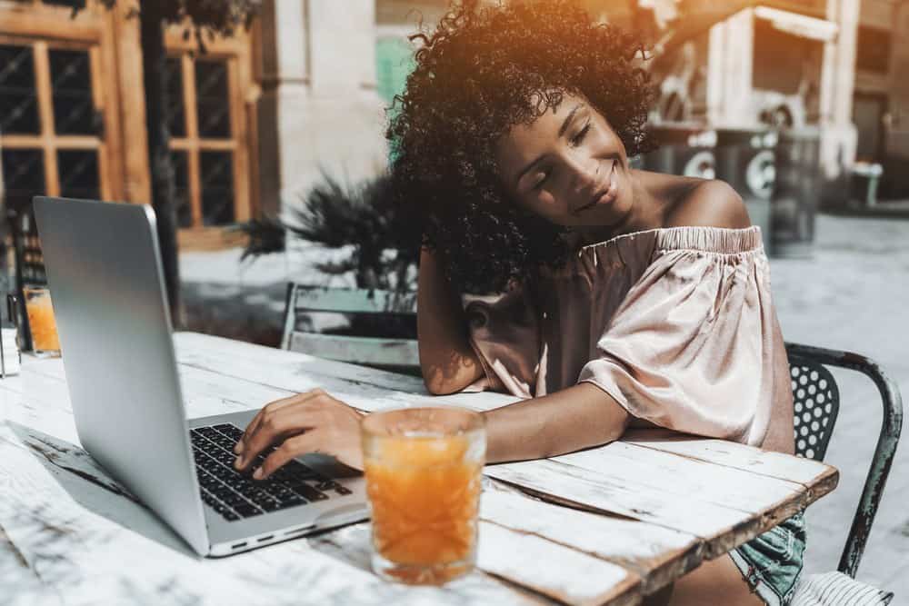 A dazzling young curly hair Brazilian female with tight ringlets admiring her hairstyle enjoying the sun while sitting in a cafe outdoors with her laptop and the glass of orange drink.