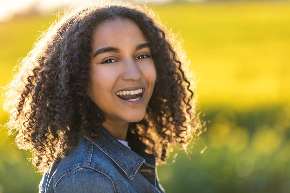 Outdoor photo of beautiful happy mixed race girl laughing with perfect teeth in field of yellow flowers, wearing a blue jean jacket with naturally curly hair.