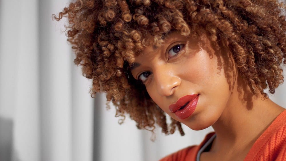 Mixed race black woman with ombre curly natural hair and natural warm make-up for dark skin  looking directly into the camera.