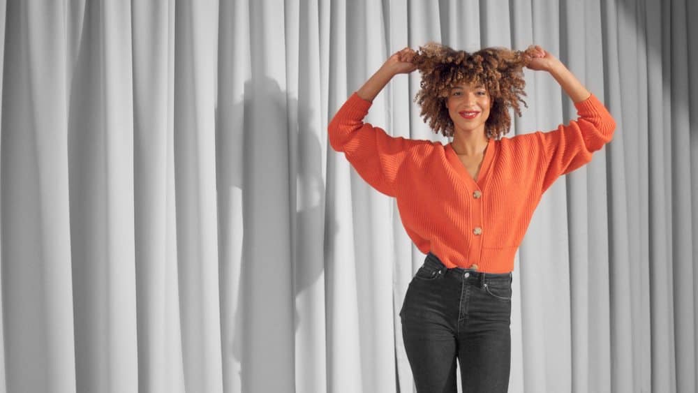 Mixed race black woman with type 3a curly hair wearing flexi rod Shirley Temple curls and natural warm make-up for dark skin dancing and touching her hair.