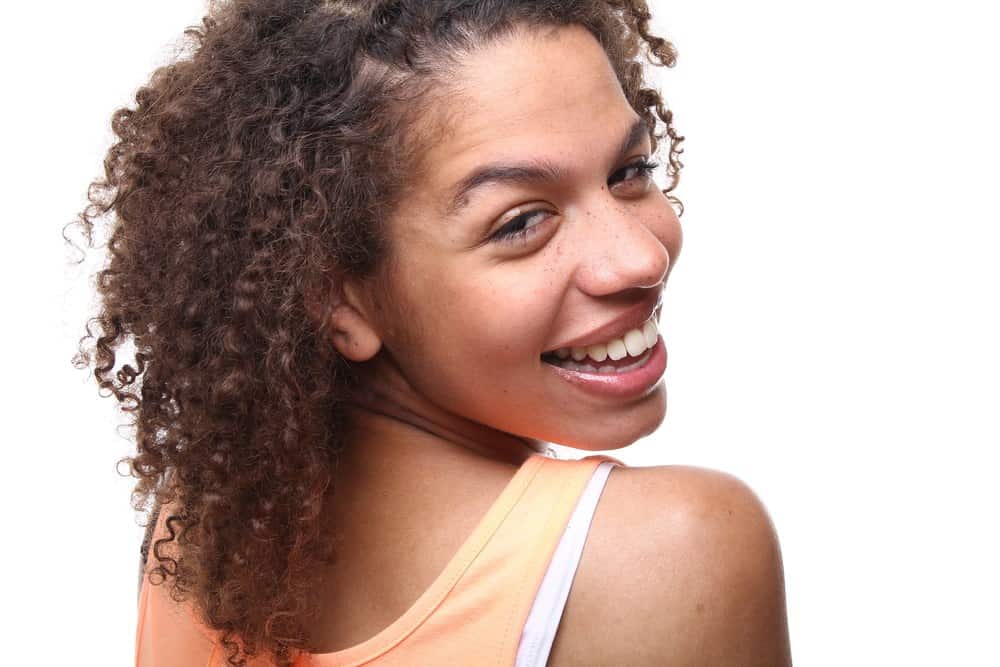 Black girl grinning looking back over her shoulder towards the camera with ombre curly type 3a hair wearing an orange tank top.