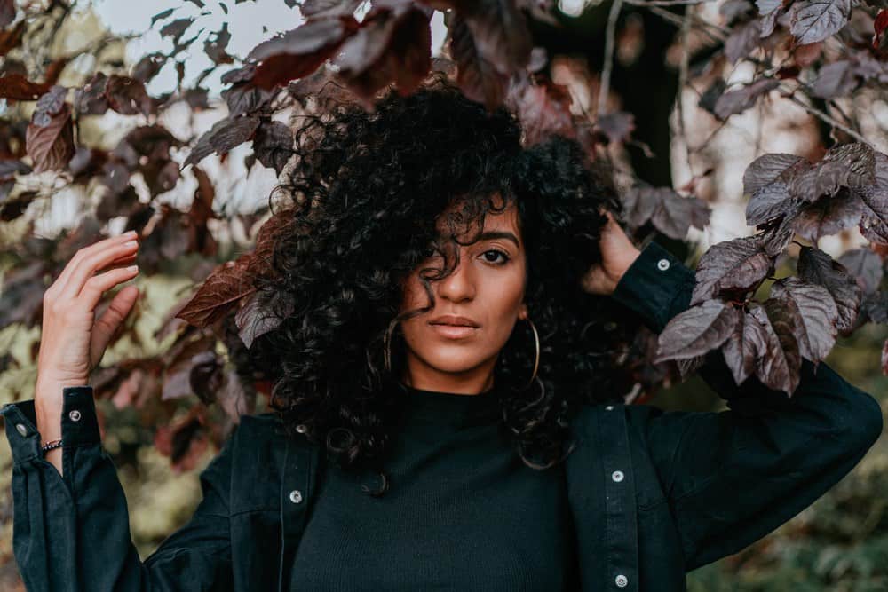 Beautiful light skinned black girl standing under a tree wearing a green jacket, green mock turtle neck shirt and gold earrings with black curly type 3 hair. 
