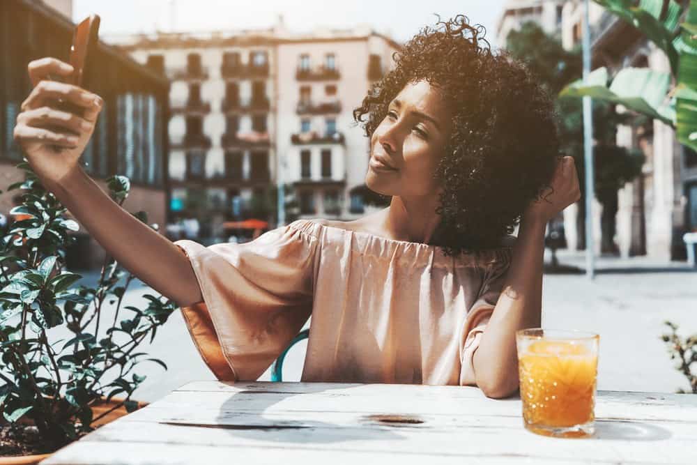 Cute biracial curly-haired girl with spiral curls taking a selfie while sitting in an outdoor bar on a sunny summer day.