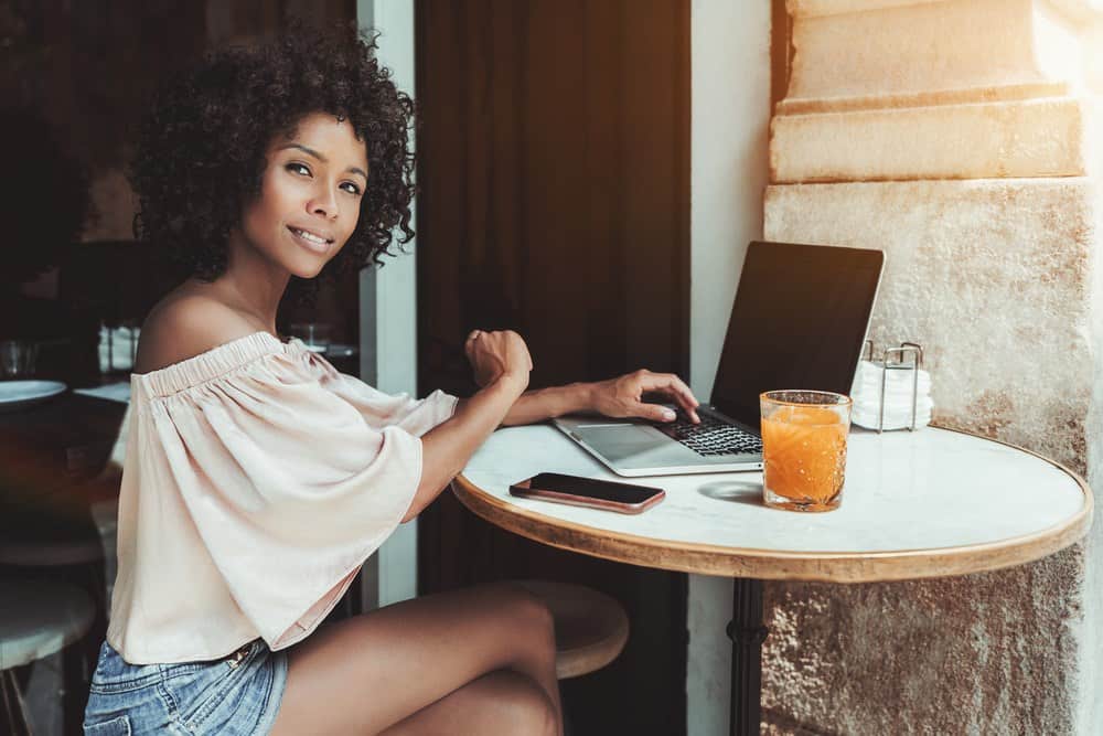 Cute African American woman with perm rods taking a picture with her iPhone while sitting at an outdoor bar on a bright autumn day.