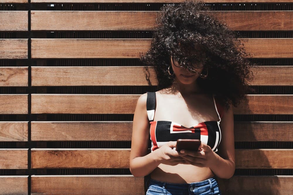 Young white woman wearing sunglasses leaning on a wooden wall using an Apple iPhone.