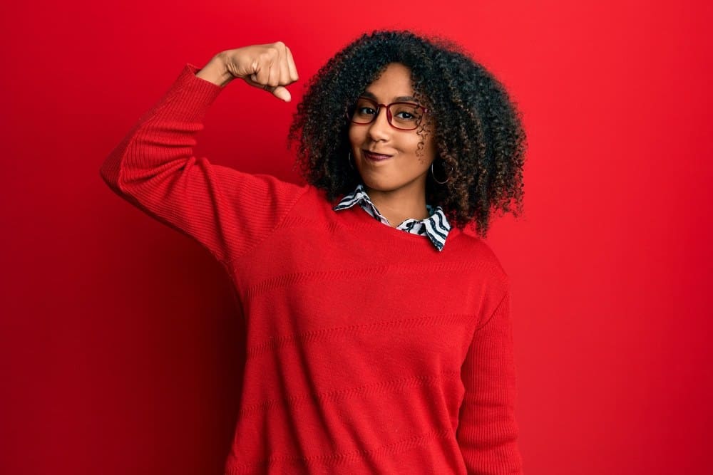 Beautiful African American woman with afro hair wearing sweater and glasses strong person showing arm muscle, confident and proud of power.