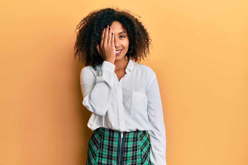 Beautiful African American woman with afro hair wearing scholar skirt covering one eye with hand, confident smile on face and surprise emotion.