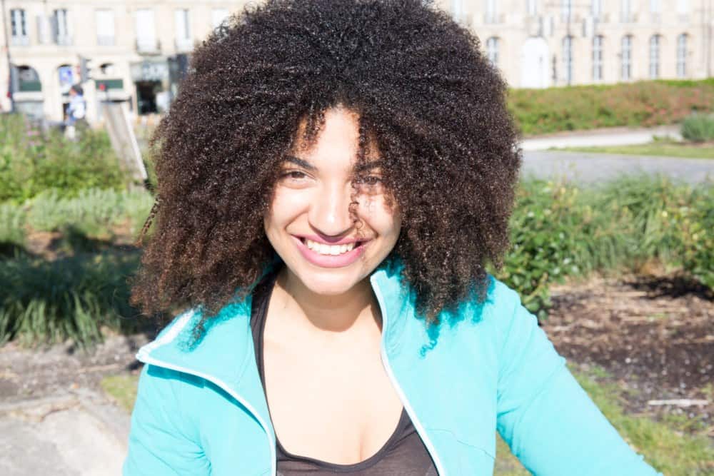 Beautiful smiling mixed girl walking in the city of Bordeaux wearing a blue a jacket, black dress, and pink lip gloss with lemongrass oil treated hair.