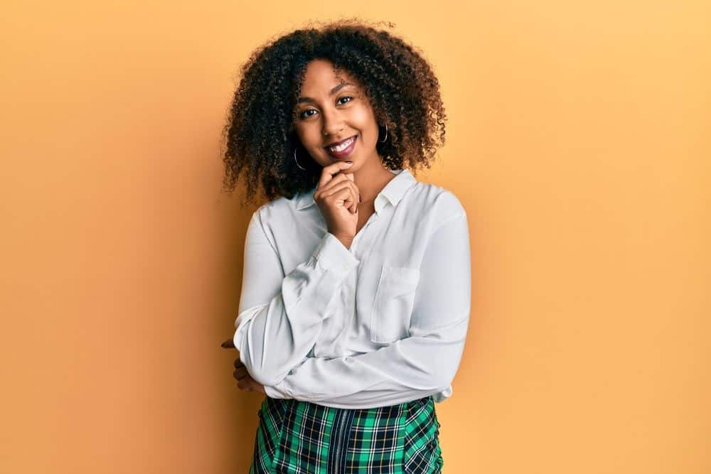 Beautiful African American woman with afro hair wearing scholar skirt looking confident at the camera with smile with crossed arms and hand raised on chin.