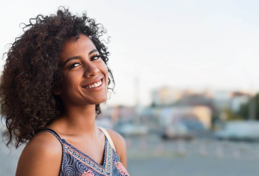 Smiling young black woman writing in her hair journal with sunlight shinning in her face wearing a dress.