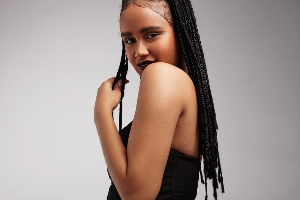 Black woman with sleek hair edges and box braids touches her braids and look directly at the camera.
