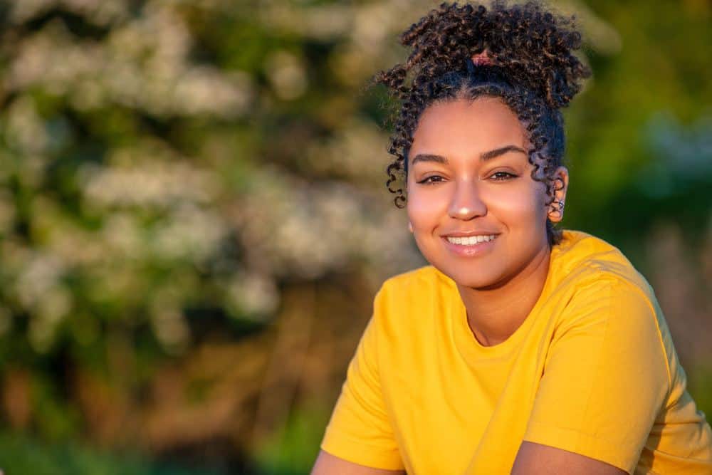 Beautiful African American young woman wearing a yellow t-shirt with curly hair, smiling with perfect teeth outside at sunset or sunrise after her hair was treated with coconut oil to protect against the UV rays outside.