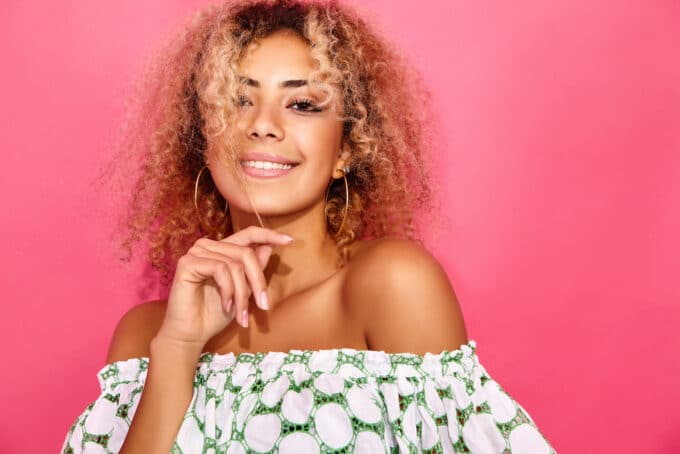 Female with ombre wavy hair wearing hoop earrings and an off-the-shoulder shirt with green and white circles.