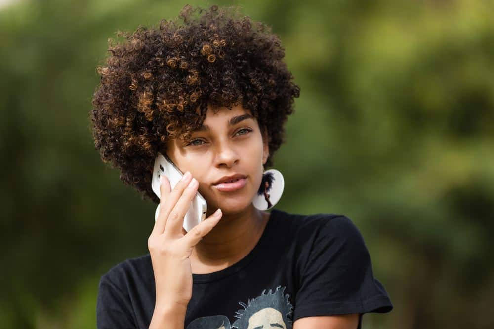 Outdoor portrait of a Young African American female speaking on mobile phone while wearing a black t-shirt and earrings featuring a lady with naturally curly hair.