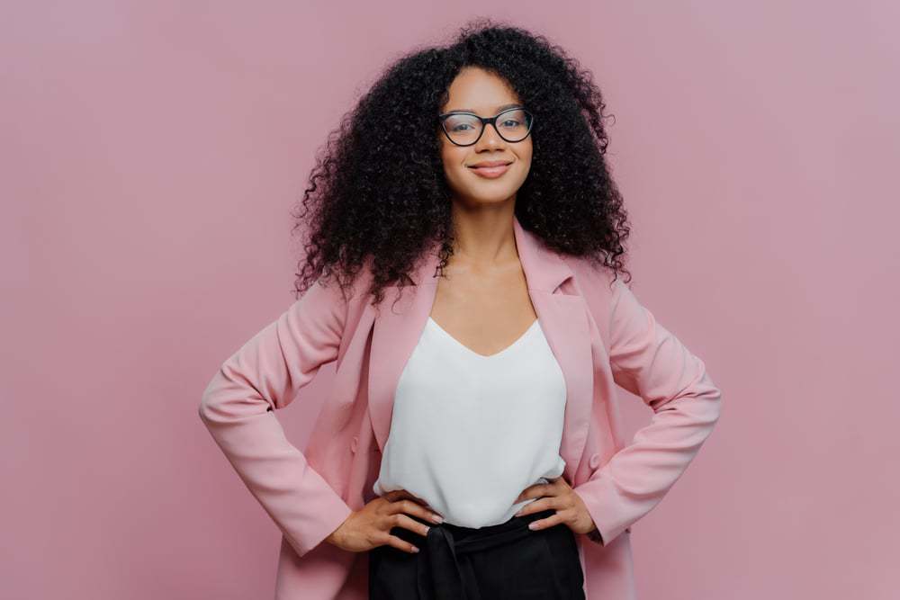 Mixed raced business woman wearing a pink blazer, white t-shirt and black glasses with black curly hair.