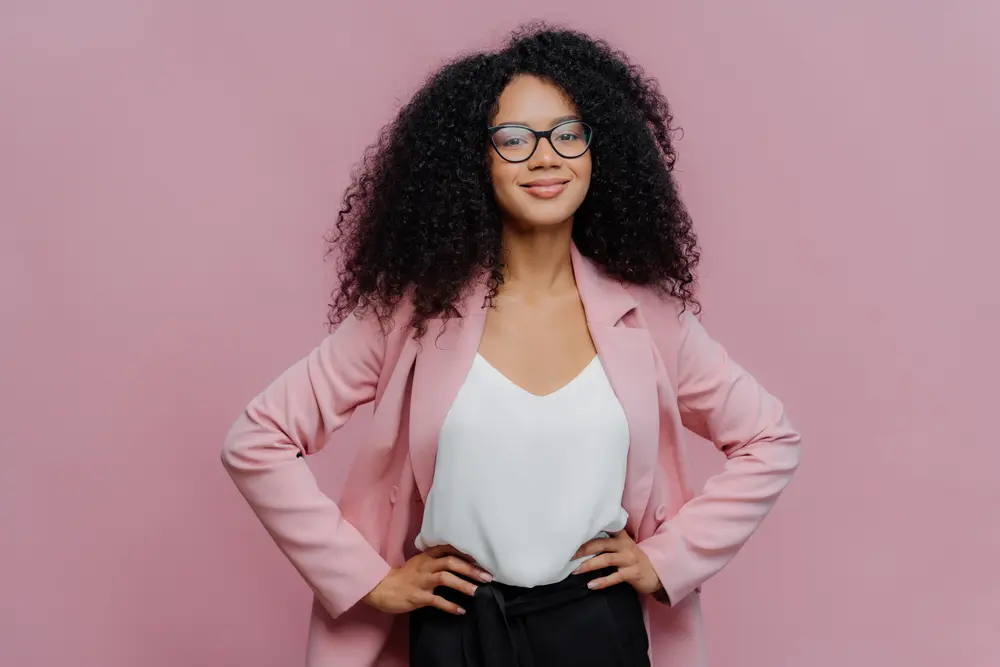 Mixed raced business woman wearing a pink blazer, white t-shirt and black glasses with black curly hair. 